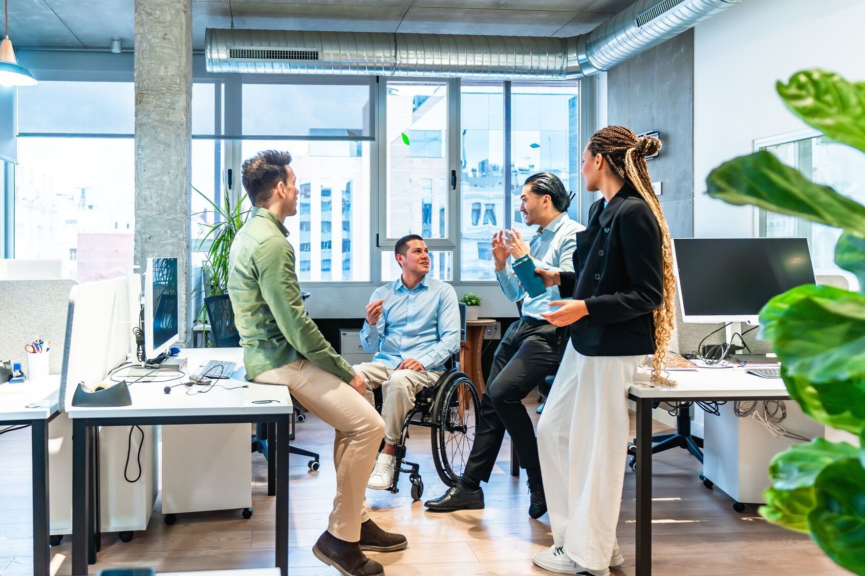 Tim bisnis Web_Wide_Diverse mendiskusikan proyek di kantor modern dengan kolega penyandang disabilitas yang menggunakan kursi roda